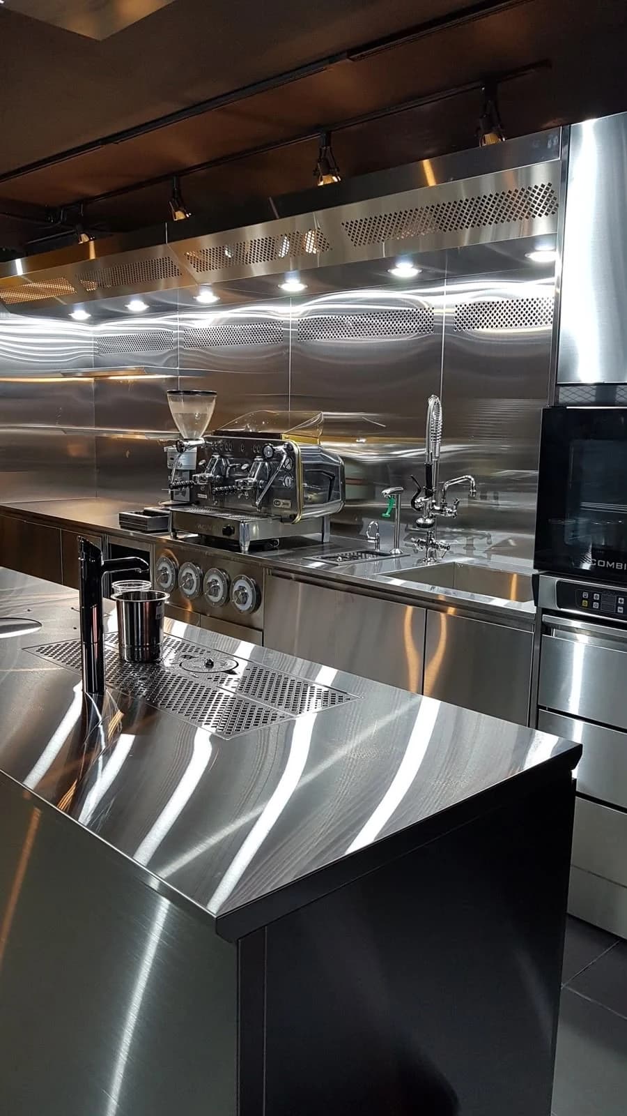 Polished stainless steel kitchen counters with coffee equipment, a sink and ventilation canopy